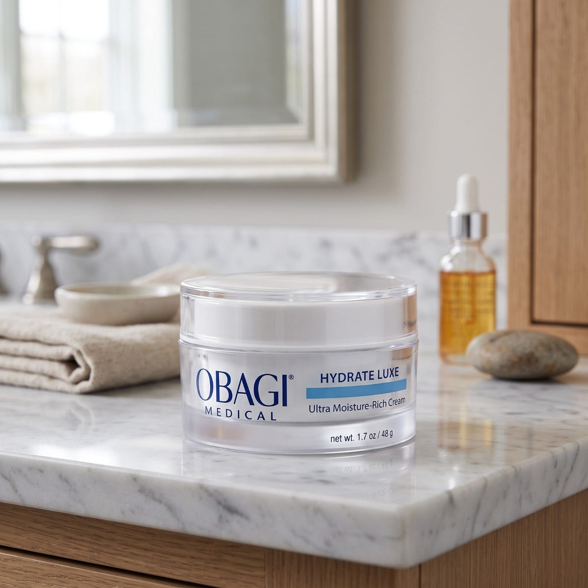 A jar of Obagi Medical Hydrate Luxe Ultra Moisture Rich Cream sits on a white marble bathroom counter next to a folded beige towel, a glass dropper bottle, and a stone, with a mirror and wooden cabinet in the background.