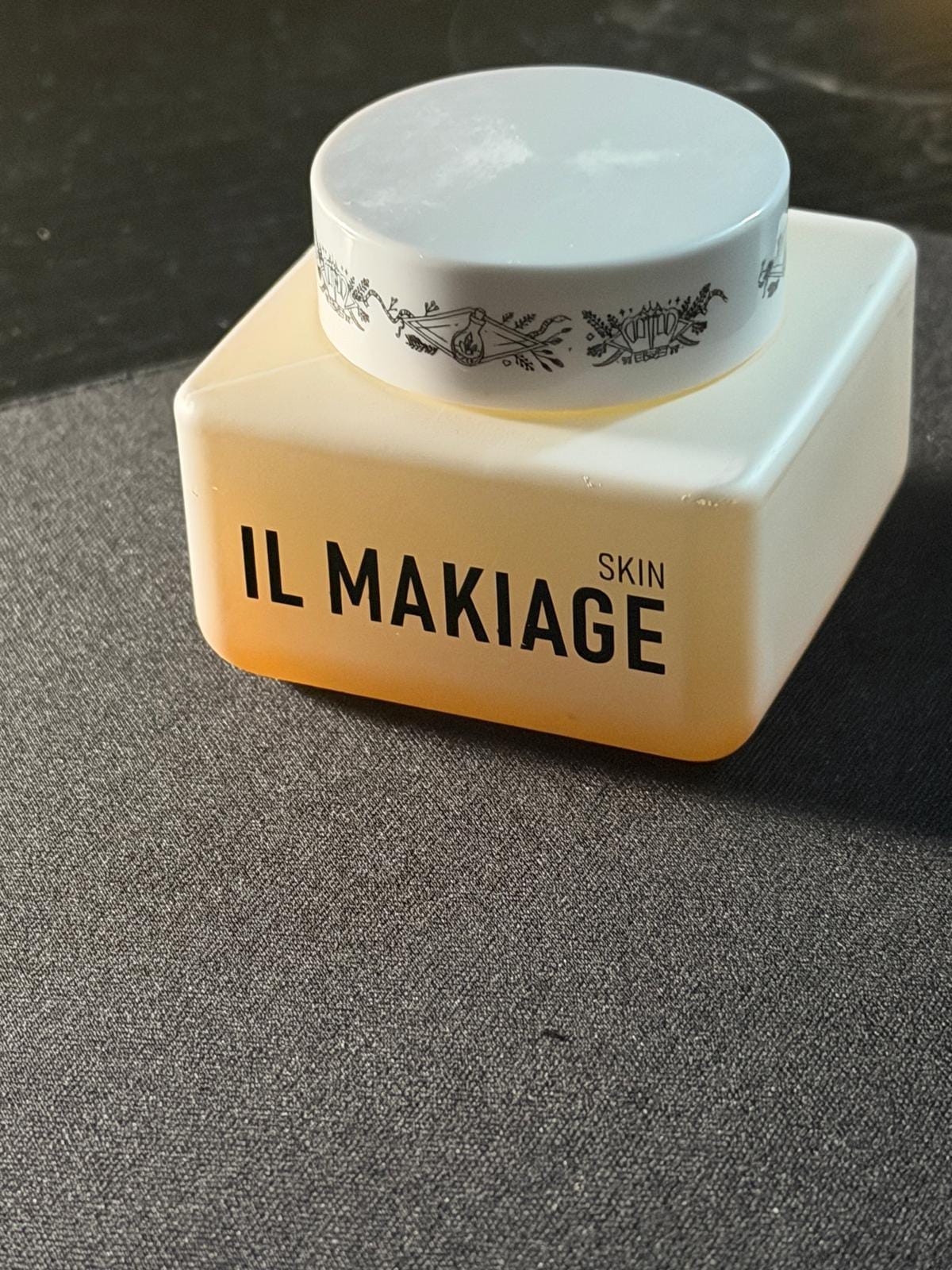 A square cream jar with a white lid and black botanical pattern, labeled 'IL MAKIAGE SKIN' in black text, resting on a dark gray textured surface.