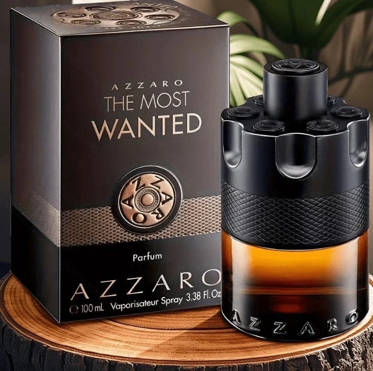 Azzaro The Most Wanted Parfum shown with its dark brown and black retail box and the perfume bottle on a wooden surface. The bottle has a black textured top and base, a clear middle section showing amber liquid, and a black textured wrap around the center.