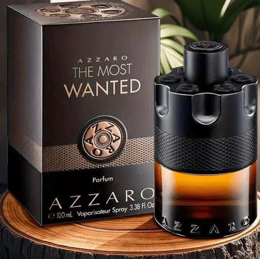 Azzaro The Most Wanted Parfum shown with its dark brown and black retail box and the perfume bottle on a wooden surface. The bottle has a black textured top and base, a clear middle section showing amber liquid, and a black textured wrap around the center.