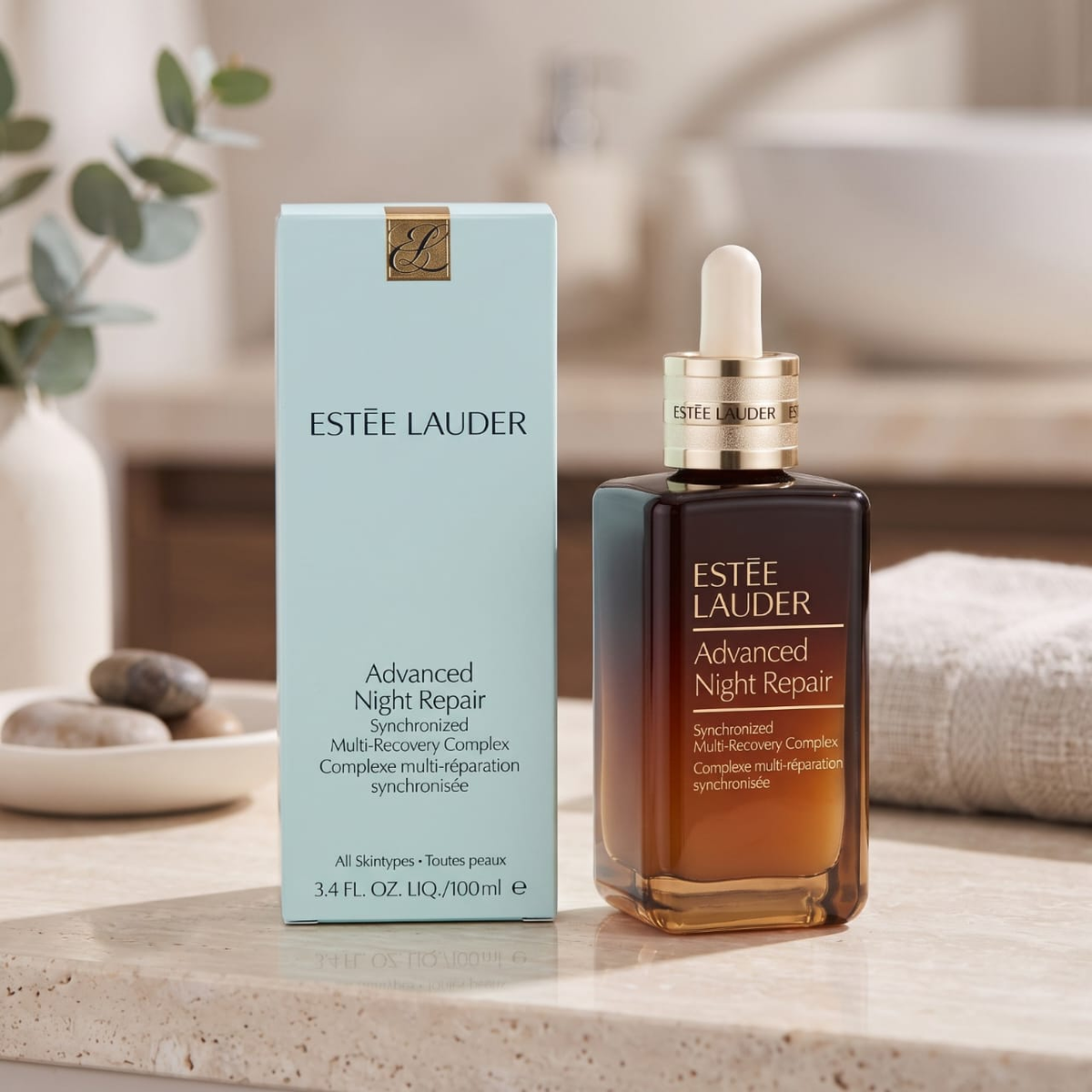 Estée Lauder Advanced Night Repair skincare set, featuring a light blue outer box and an amber glass dropper bottle with a gold collar and white dropper cap, displayed on a marble countertop with a softly blurred bathroom background.