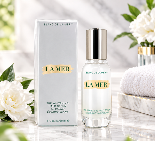 La Mer Blanc de La Mer The Whitening Halo Serum shown as a clear glass bottle with a silver cap next to its white box packaging, styled on a marble surface with white flowers and a towel in the background.