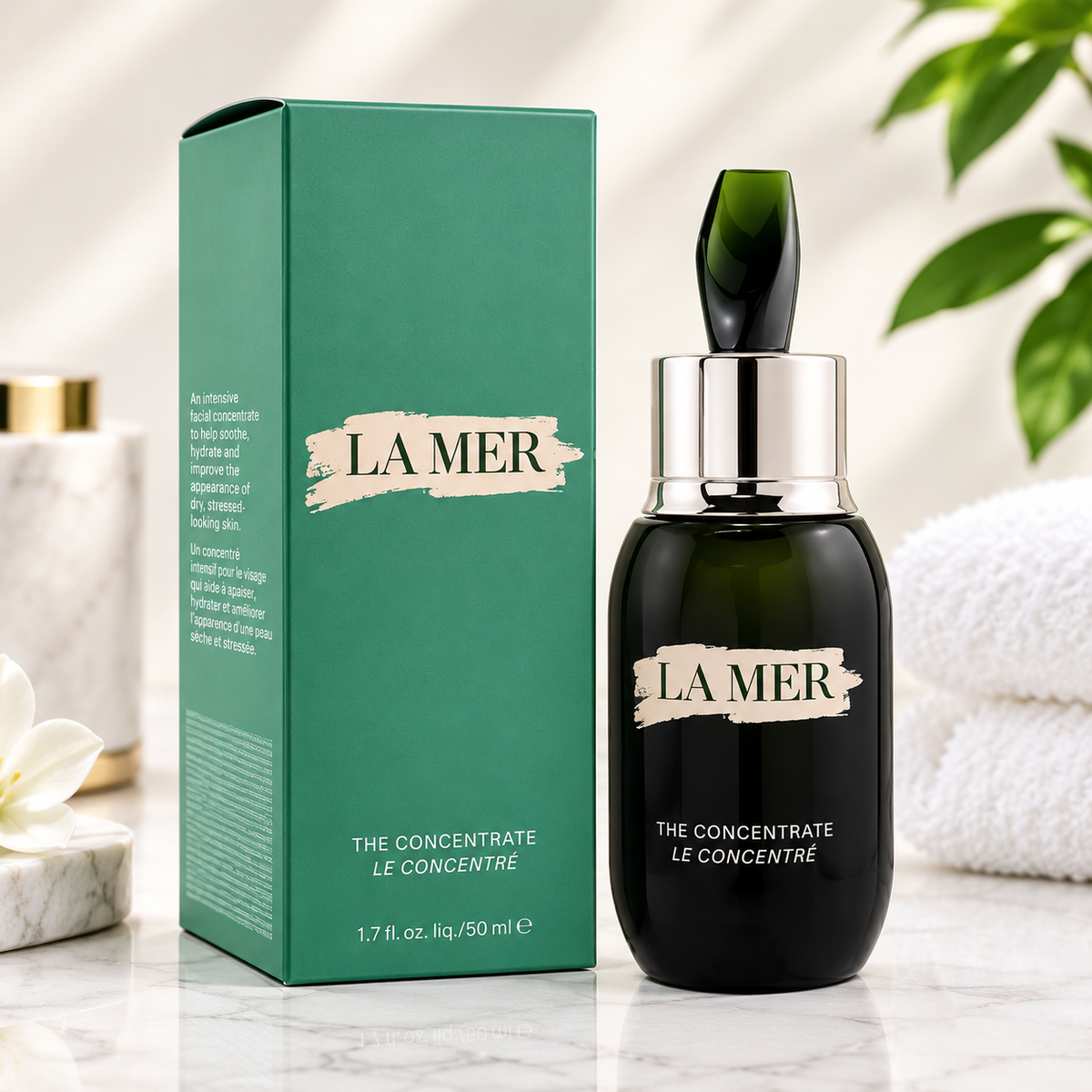 La Mer The Concentrate Le Concentré skincare set featuring a dark green glass dropper bottle and its matching green packaging box, styled on a marble surface with a white towel and green leaves in the background.