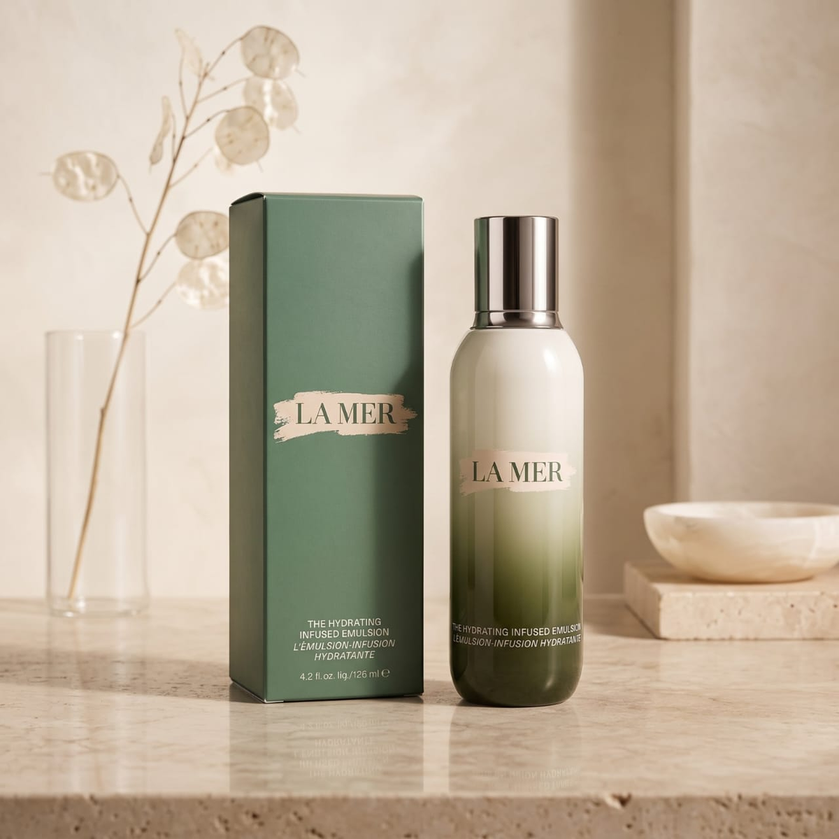 La Mer Hydrating Infused Balmy Milk bottle with a silver cap next to its green packaging box, displayed on a beige stone surface with neutral decorative props.