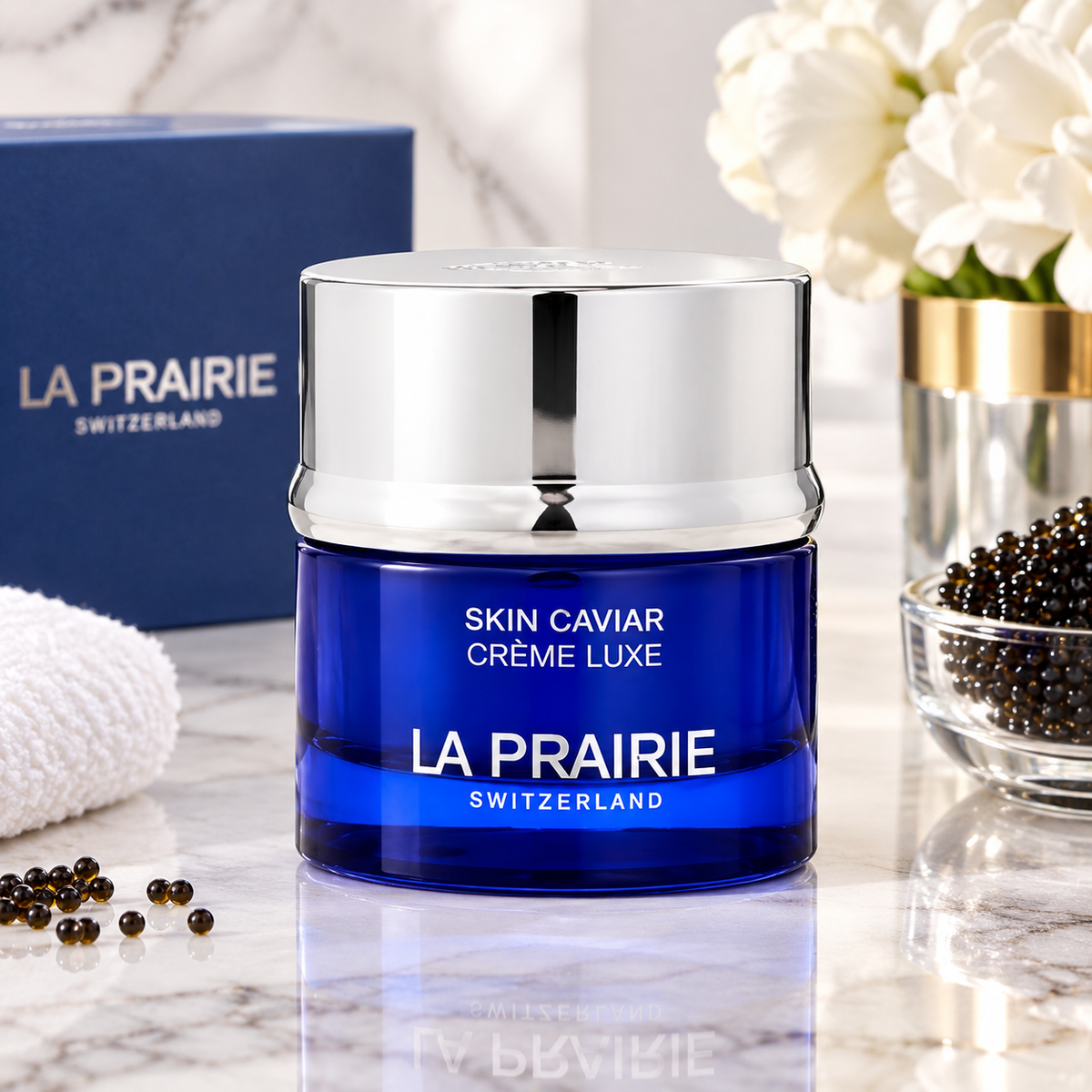 A jar of La Prairie Skin Caviar Crème Luxe in a deep blue glass jar with a reflective silver lid, set on a marble surface with its blue box, white flowers, and caviar visible in the background.