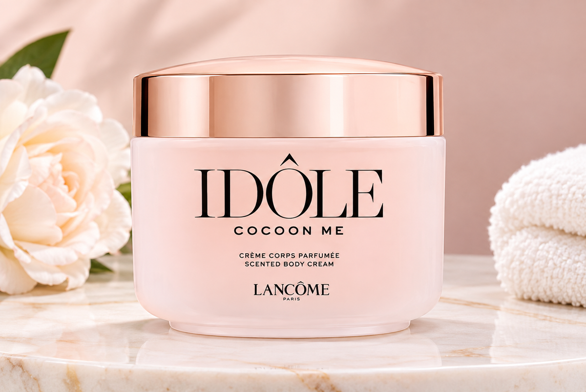 Lancôme Idôle Cocoon Me scented body cream in a frosted pale pink jar with a metallic rose gold lid, displayed on a marble surface with a white rose and folded white towels in the background.