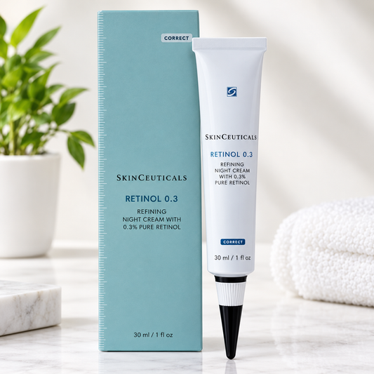 SkinCeuticals Retinol 0.3 retining night cream in a white squeeze tube with a black applicator, standing next to its light blue box on a marble surface with a blurred plant and towel in the background.