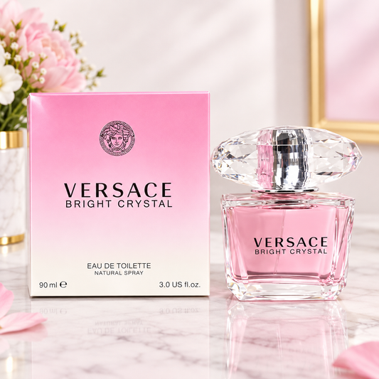 Versace Bright Crystal Eau de Toilette perfume bottle and its pink gradient box, displayed on a marble surface with soft pink flowers in the background.