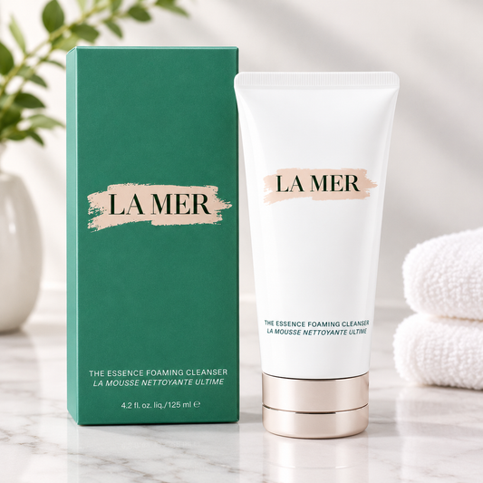 La Mer The Essence Foaming Cleanser shown with its green box and white squeeze tube on a marble surface, with a soft blurred background including a plant and folded white towels.